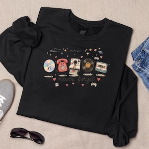 May include: A black long-sleeve sweatshirt featuring a graphic print of retro technology items including a CD, rotary phone, instant camera, vinyl record, and cassette tape with the text "never forget" surrounded by small hearts and stars.