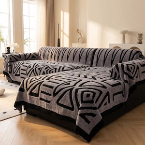 May include: A gray and black textured sofa cover featuring a repeating geometric pattern of triangles, squares, and parallel lines. The fabric has a plush, soft appearance and drapes over the entire surface of the sectional sofa, covering the back, seat, and sides.