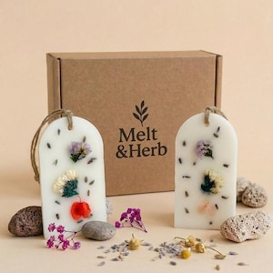 Könnte beinhalten: Zwei weiße, bogenförmige Wachstafeln mit eingebetteten Trockenblumen und Lavendelknospen, jeweils mit einer Juteschnur zum Aufhängen. Dahinter steht ein brauner Karton mit dem Aufdruck Melt & Herb und einem Blattsymbol.