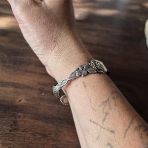Könnte beinhalten: Ein silberfarbener Metall-Armreif mit geprägtem Blumenmuster, detaillierten Blättern und einem Rosenmotiv. Das Armband hat eine offene Struktur mit einem strukturierten, rankenartigen Muster, das vom zentralen Blumenelement ausgeht.