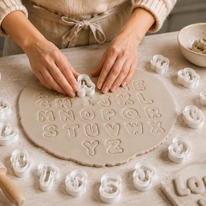 May include: A set of white plastic alphabet cookie cutters used to press letters into a flat, circular piece of light beige dough. The cutters are arranged around the dough, which features the full alphabet embossed into its surface.