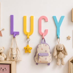 Könnte beinhalten: Vier große, bunte Wandhaken in Buchstabenform, die das Wort LUCY in Lila, Gelb, Rosa und Blau bilden. Jeder Buchstabe hat unten einen Haken, an dem eine beige Tragetasche, ein gelber Stern-Schlüsselanhänger, ein Blumenrucksack und ein Stoffhase hängen.