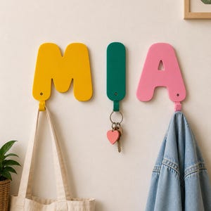 Op de afbeelding: Drie wandhaken in de vorm van de letters M, I en A. De M is geel, de I is een donkergroene verticale balk en de A is roze. Elke letter heeft onderaan een klein haakje. Een draagtas hangt aan de M, sleutels aan de I en een spijkerjas aan de A.