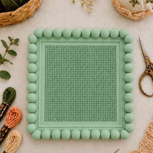 May include: A rectangular sage green embroidery frame featuring a border of uniform spherical beads. The center of the frame contains a fine, grid-like mesh surface designed for needlework projects, providing a structured base for stitching.