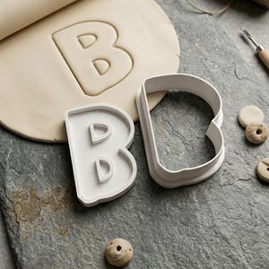 Puede incluir: Dos cortadores de galletas de plástico blanco con forma de letra B, uno con un sello interno para detalles, junto a una lámina de arcilla beige extendida con una impresión de la letra B y varias cuentas de arcilla pequeñas y texturizadas.