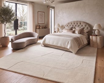 Puede incluir: Una alfombra rectangular grande de color crema neutro con un sutil patrón geométrico de líneas y semicírculos tallados. La alfombra está colocada sobre un suelo de madera en un dormitorio, cubriendo una parte importante de la superficie.