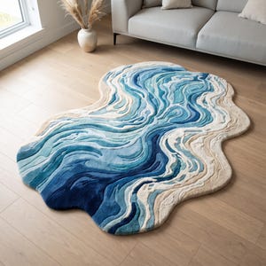 Puede incluir: Una alfombra de forma irregular con un dise&ntilde;o texturizado y en capas de remolinos en tonos azul, turquesa y crema que imitan la apariencia de las olas del oc&eacute;ano y las costas arenosas, con una estructura de pelo alto y bajo y bordes fluidos.
