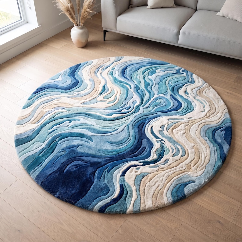 Puede incluir: Una alfombra circular con un patr&oacute;n abstracto y texturizado de olas arremolinadas en tonos azul marino, azul claro, blanco y beige. El dise&ntilde;o imita el agua que fluye con secciones elevadas y talladas que crean profundidad en la superficie.