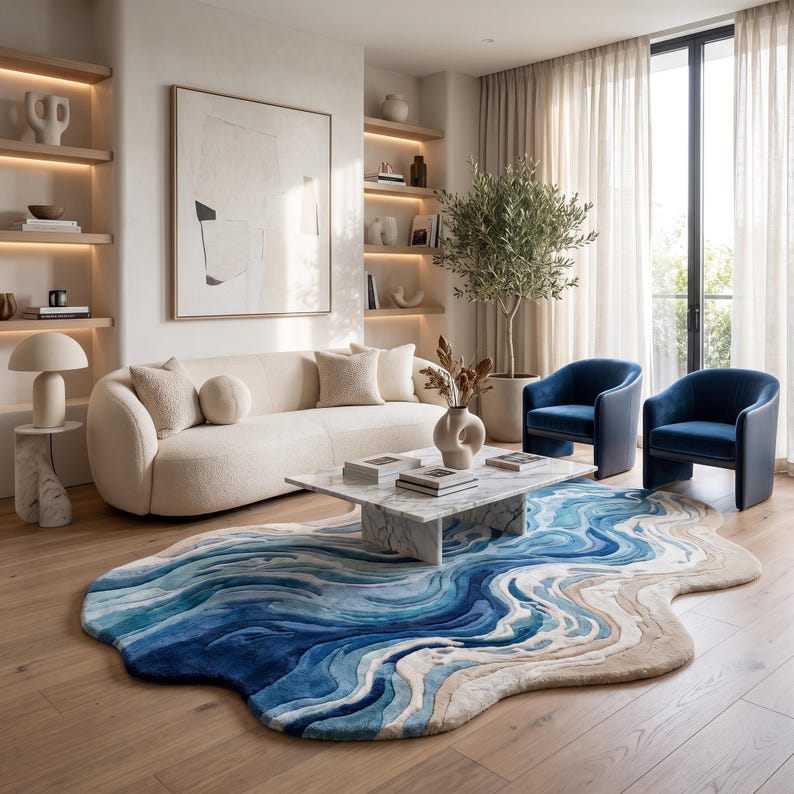 Puede incluir: Una alfombra de forma org&aacute;nica irregular con un patr&oacute;n fluido y superpuesto en tonos azul profundo, azul claro y beige arena. La alfombra est&aacute; colocada sobre un suelo de madera clara bajo una mesa de centro de m&aacute;rmol blanco en una sala.