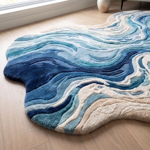 Puede incluir: Una alfombra de forma irregular con una superficie texturizada y copetuda que presenta un patr&oacute;n abstracto y arremolinado en tonos azul marino profundo, turquesa brillante, azul cielo claro, crema y beige, imitando el aspecto del agua.