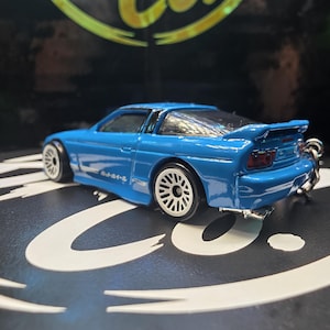 May include: A bright blue die-cast toy car featuring white multi-spoke wheels, a rear spoiler, and Japanese text printed on the side panel. The vehicle includes a metal keychain attachment at the rear and has red taillight details.