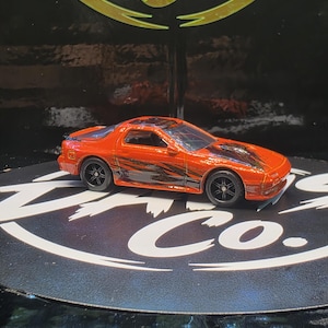 May include: A die-cast orange sports car toy featuring black racing stripes along the sides and hood, black wheels, and a black rear spoiler. The vehicle has a sleek, low-profile design with tinted windows and a detailed exterior finish.