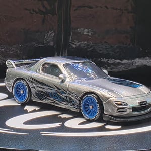 May include: A silver die-cast toy car featuring a black and blue graphic design along the sides, bright blue multi-spoke wheels with white lettering on the tires, a rear spoiler, and a front license plate clearly displaying the text RX-7.