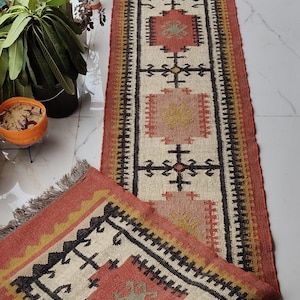 May include: A long, rectangular runner rug featuring a cream-coloured centre with repeating geometric patterns in terracotta, black, and mustard yellow. The rug has a wide terracotta border, a textured woven surface, and fringed ends on both sides.