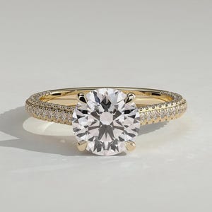 Könnte beinhalten: Ein Ring aus Gelbgold mit einem großen, runden, im Brillantschliff geschliffenen, klaren Mittelstein, der von vier Krappen gehalten wird. Die Ringschiene ist mit kleinen, klaren, rund geschliffenen Steinen in Pavé-Fassung besetzt.