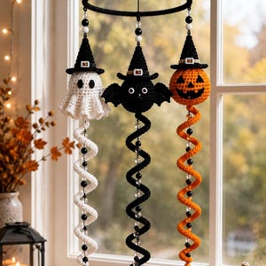 May include: A hanging mobile featuring three crocheted figures: a white ghost, a black bat, and an orange jack-o'-lantern, each wearing a black witch hat. Below each figure hangs a long, spiraling crocheted tail accented with clear and black beads.