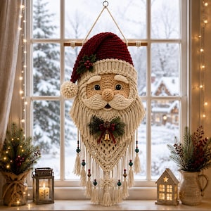 Könnte beinhalten: Ein Makramee-Wandbehang in Form eines Weihnachtsmann-Gesichts mit einer dunkelroten Häkelmütze samt weißem Bommel, einem cremefarbenen Bart mit Fransen, einer kleinen goldenen Glocke, grünen Stechpalmenblättern und roten Perlen am unteren Rand.