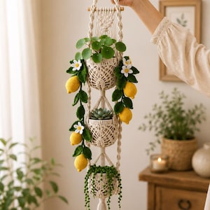 May include: A vertical macrame plant hanger featuring three cream-colored crocheted baskets. The structure is adorned with crocheted green leaves, white flowers, and yellow lemons. It holds a pilea plant, a succulent, and a string of pearls plant.