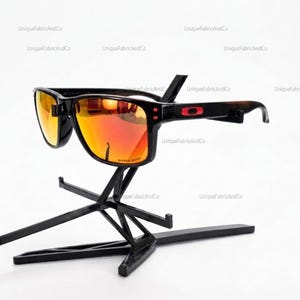 Pode incluir: Óculos de sol pretos com armação retangular e lentes laranja refletoras. As hastes apresentam um logótipo vermelho. Os óculos estão expostos num suporte de plástico preto. A palavra Prizm está escrita no canto inferior da lente.