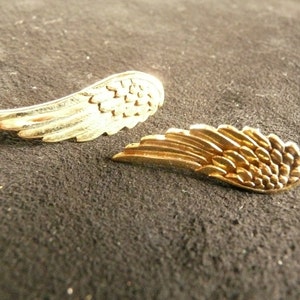 May include: Two gold-colored angel wing earrings. The wings are detailed with a textured pattern.
