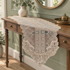 Puede incluir: Un camino de mesa de encaje beige con un intrincado patrón de bordado de flores y enredaderas con bordes festoneados. El camino está hecho de una tela fina y transparente y muestra un diseño simétrico que recorre el centro y termina en puntas.