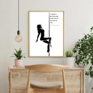 May include: A framed wall art print featuring a black and white silhouette of a person on a pole. The text reads: IT DOESN'T MATTER HOW SLOW YOU GO AS LONG AS YOU.. never stop... The print is displayed in a light-coloured wooden frame on a wall.