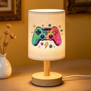 May include: A cylindrical table lamp with a light-coloured fabric shade featuring a colourful, multi-hued video game controller graphic. The design includes small stars, circles, and a lightning bolt icon. The lamp has a light wood base and stem.