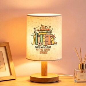 May include: A cylindrical table lamp with a light-colored fabric shade featuring a colorful illustration of stacked books, flowers, and stars. The text "THERE IS NO SUCH THING AS TOO MANY BOOKS" is printed below the books. It has a wooden base.