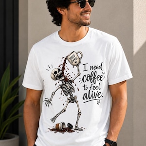Può includere: Una t-shirt bianca con la grafica di uno scheletro che versa del caffè da una tazza sulla propria testa. Il testo recita "I need coffee to feel alive." con un piccolo simbolo a forma di cuore. La tazza riporta la scritta "NEED COFFEE" con un cuore.