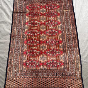 May include: A rectangular area rug featuring a repeating geometric pattern of diamonds and octagons in shades of red, orange, and cream. The rug is framed by a dark blue border with intricate linear motifs and finished with white fringe at both ends.