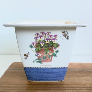 May include: A white ceramic planter with a flared rim, featuring a central illustration of purple and yellow Viola tricolor flowers in a terracotta pot, accompanied by two butterflies and a blue crackle-patterned base.
