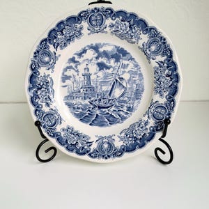 May include: A white ceramic plate featuring a blue transferware pattern. The centre depicts a lighthouse and a sailboat on choppy water. The rim is decorated with a detailed border of floral motifs and small crests, displayed on a black metal stand.