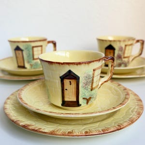 May include: A set of three pale yellow ceramic tea cups and matching saucers. Each cup features a hand-painted design of a small cottage with a brown door, a window, and green foliage. The saucers have a textured, embossed stone-like pattern.
