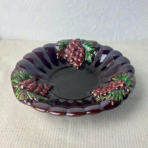 May include: A round, dark brown ceramic bowl with a scalloped edge and a glossy finish. The bowl features three raised, embossed clusters of purple grapes with green leaves arranged symmetrically around the interior rim of the dish.