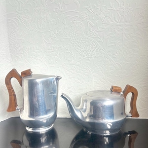 May include: A silver-coloured metal pitcher and a matching teapot, both featuring polished metallic bodies and distinctively shaped wooden handles with small wooden lid knobs, displayed side by side on a reflective dark surface.