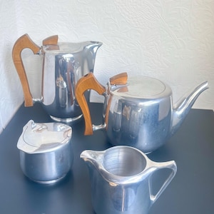 May include: A four-piece tea and coffee service set made of polished silver-toned metal. The set includes a tall coffee pot, a teapot, a lidded sugar bowl, and a creamer. The coffee pot and teapot feature distinct light brown wooden handles.