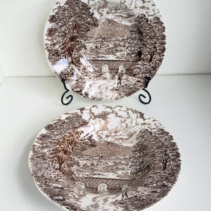 May include: Two white ceramic plates featuring a brown transferware pattern of a rural landscape. The design depicts a stone bridge over a stream, trees, and a distant village with a church spire. One plate is upright on a black metal stand.