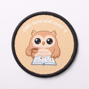 May include: A circular embroidered patch with a beige background and a black stitched border. The design features a brown owl with large black eyes, holding a steaming brown mug and reading an open book. The text reads OWL-lone and lovin' it.