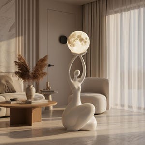 May include: A white floor lamp featuring a stylised human figure in a seated pose, holding a spherical, glowing moon-textured light above its head. The lamp has a smooth, matte finish and emits a warm, soft light from the circular orb.