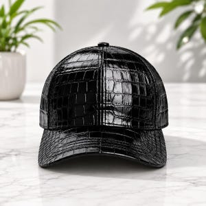 Pode incluir: Um boné de beisebol preto com acabamento brilhante e envernizado, apresentando uma textura de pele de crocodilo. O boné possui uma copa estruturada e uma aba curva, exibindo uma superfície reflexiva que destaca os detalhes em relevo.
