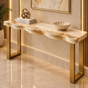 Può includere: Un tavolo console con un piano in marmo bianco e oro dai bordi ondulati, sostenuto da due gambe rettangolari in metallo dorato. Sulla superficie sono appoggiati due libri intitolati The Kinfolk Home e Louis Vuitton, una scultura a nodo dorata e una ciotola bianca.