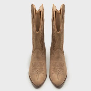 Op de afbeelding: Een paar beige leren cowboylaarzen met een spitse neus, een schacht tot halverwege de kuit met aantreklussen en ingewikkelde decoratieve stiksels op de wreef en de schacht. De laarzen hebben een blokhak en een gladde, matte afwerking.