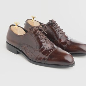 May include: A pair of dark brown leather oxford shoes with a polished finish and a textured crocodile-embossed pattern on the side panels. The shoes feature matching brown laces, a rounded toe, a black sole, and light-coloured wooden shoe trees.