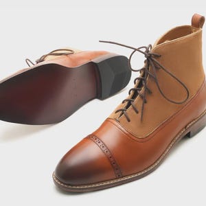 May include: A pair of lace-up ankle boots featuring a tan suede upper and a polished brown leather toe cap with decorative brogue detailing. The boots include dark brown laces, a pull tab at the heel, and a smooth dark brown leather sole.