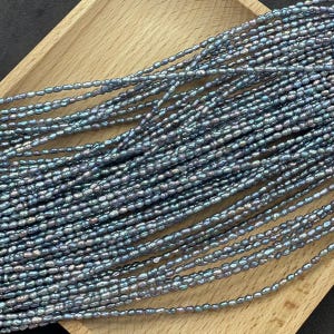 Puede incluir: Una colección de pequeñas perlas irregulares de color azul oscuro y gris iridiscente, ensartadas en múltiples hilos finos. Las perlas presentan un brillo metálico con sutiles tonos violetas y verdes, creando una apariencia texturizada y brillante.