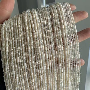 May include: A collection of multiple strands of tiny, off-white seed pearls arranged in parallel lines. Each strand consists of numerous small, round, textured pearls with a subtle lustre, creating a dense and uniform appearance across the group.