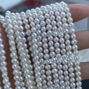 May include: Multiple strands of small, round, white freshwater pearls arranged in parallel lines. Each pearl features a smooth, lustrous surface with subtle variations in shape and size, reflecting light across the entire collection of beads.