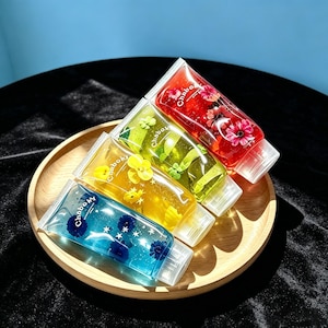 Può includere: Quattro tubetti trasparenti riempiti di gel colorato e fiori incorporati. I tubetti sono rossi, verdi, gialli e blu, ognuno con il marchio Chaboky stampato in bianco insieme a piccoli accenti a forma di stella e dettagli floreali.