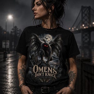 Puede incluir: Una camiseta negra con un gráfico de una criatura alada con ojos rojos brillantes, una luna llena y un puente. El texto dice "OMENS DON'T KNOCK" sobre "POINT PLEASANT WEST VIRGINIA". El diseño incluye ilustraciones de polillas y un bosque oscuro.