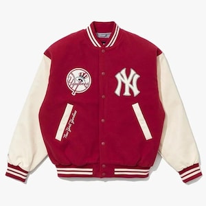 Op de afbeelding: Een rood varsity-jack met crèmekleurige mouwen, voorzien van een wit New York Yankees-logo op de borst, een ronde honkbalpatch met een hoge hoed en de tekst New York Yankees geborduurd in wit schrift bij de rechterzak.